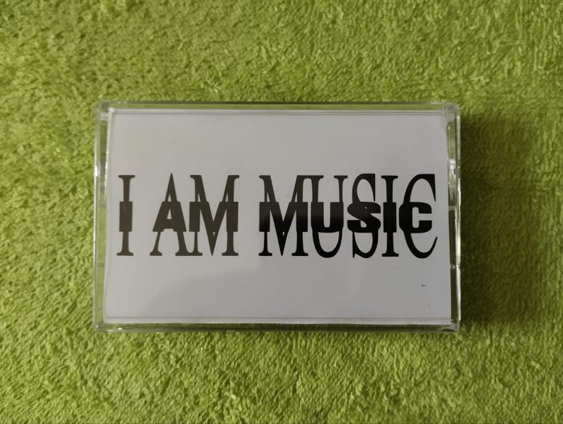 May include: A clear cassette tape case with a white insert. The insert features the text "I AM MUSIC" in a bold, black font. The background is a textured, green surface.
