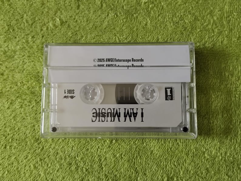 May include: A clear cassette tape with the words "I AM MUSIC" printed on the label. The tape is in a clear plastic case, and the background is a green surface. The text "SIDE 1" is also visible.