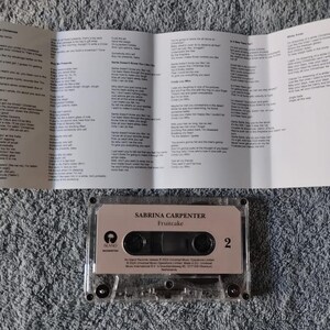 May include: A clear cassette tape case with a pink label that reads "Sabrina Carpenter -  "Fruitcake"  in white text. The case is sitting on a grey, fuzzy surface.