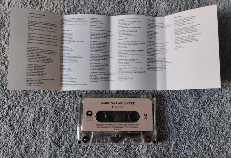 May include: A clear cassette tape case with a pink label that reads "Sabrina Carpenter -  "Fruitcake"  in white text. The case is sitting on a grey, fuzzy surface.