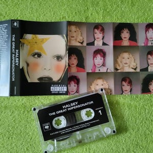 May include: A cassette tape and album art for Halsey's "The Great Impersonator." The cassette tape is clear with black text. The album art features a close-up of a face with a yellow star and multiple images of the artist.