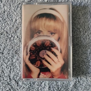 May include: A cassette tape featuring a photo of a woman holding a plate of pecan pie. The woman is wearing a white headband and a red sweater with a heart design. The text "118" is visible on her hand.