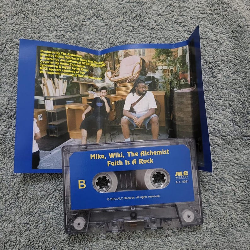 May include: A blue cassette tape with the text "Mike, Wiki, The Alchemist Faith Is A Rock" and "ALC Records ALC-5051" printed on it. The tape is surrounded by a blue background with a photo of two men sitting on a bench.