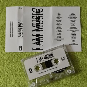 May include: A clear cassette tape labeled "I AM MUSIC" with a tracklist on a white insert. The cassette is on a green surface. The insert lists song titles and artist names.