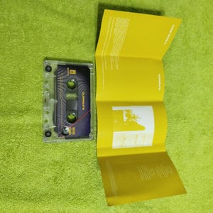 May include: A transparent cassette tape with a black and blue label, displaying the word "CURRENTS" and "SIDE TWO". A yellow booklet is open beside the cassette. The background is a solid green colour.