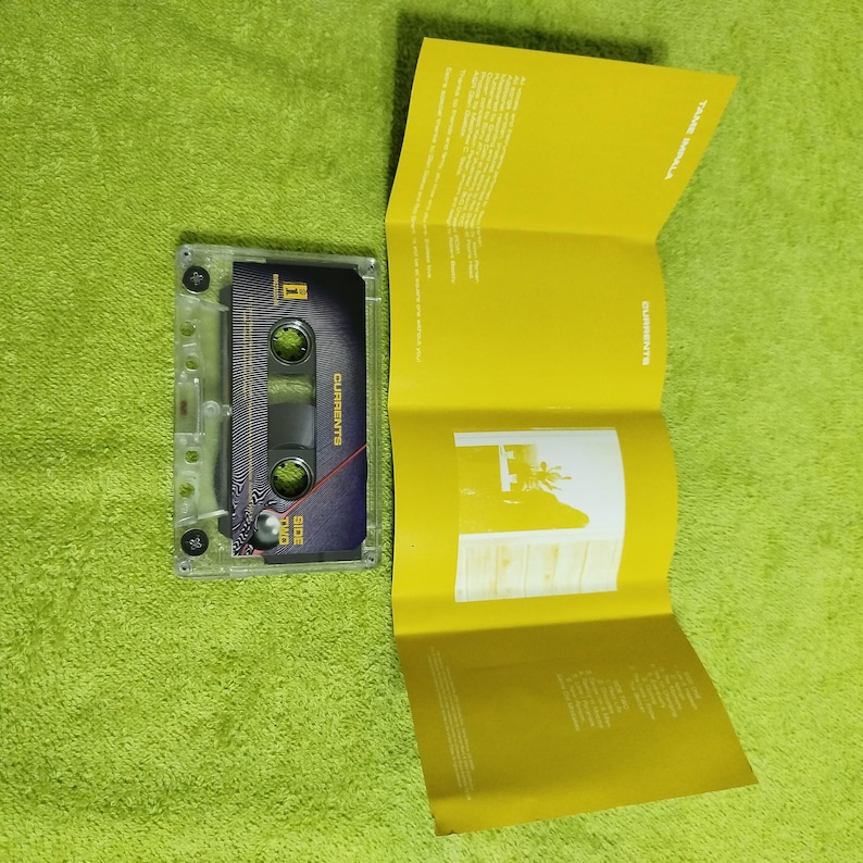May include: A transparent cassette tape with a black and blue label, displaying the word "CURRENTS" and "SIDE TWO". A yellow booklet is open beside the cassette. The background is a solid green colour.