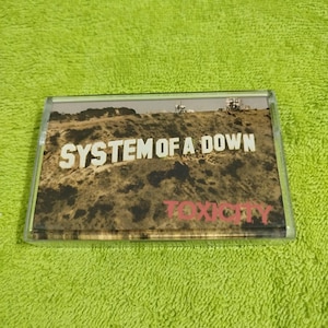 May include: A cassette tape with a brown and green landscape image. The text "SYSTEM OF A DOWN" is printed in white on the tape. The text "TOXICITY" is printed in red on the tape.