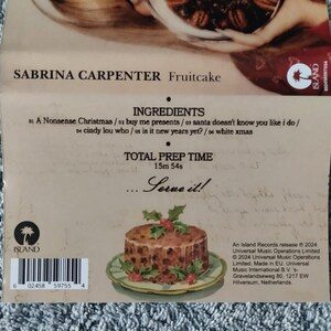 May include: A CD case for the album "Fruitcake" by Sabrina Carpenter. The case features a picture of a fruitcake with green holly leaves on top. The text "Serve it!" is printed on the case, along with the album title and artist name. The back of the case includes a barcode and information about the record label and release date.