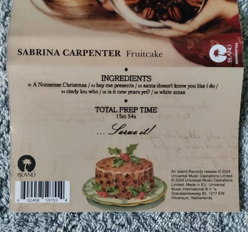 May include: A CD case for the album "Fruitcake" by Sabrina Carpenter. The case features a picture of a fruitcake with green holly leaves on top. The text "Serve it!" is printed on the case, along with the album title and artist name. The back of the case includes a barcode and information about the record label and release date.