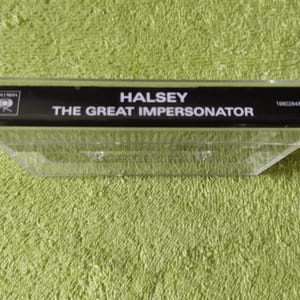 May include: A clear plastic cassette tape case with a black label. The label reads "HALSEY THE GREAT IMPERSONATOR" in white text. The Columbia Records logo is visible. The case is on a green textured surface.