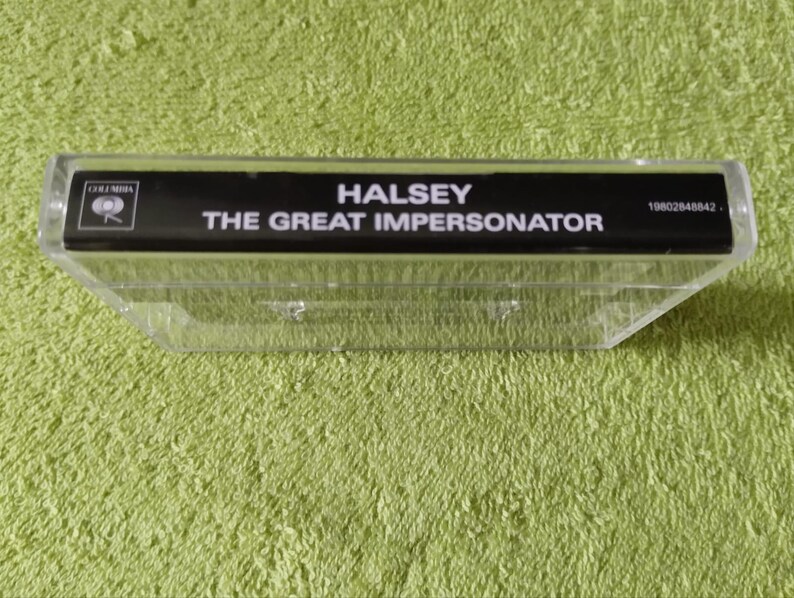 May include: A clear plastic cassette tape case with a black label. The label reads "HALSEY THE GREAT IMPERSONATOR" in white text. The Columbia Records logo is visible. The case is on a green textured surface.