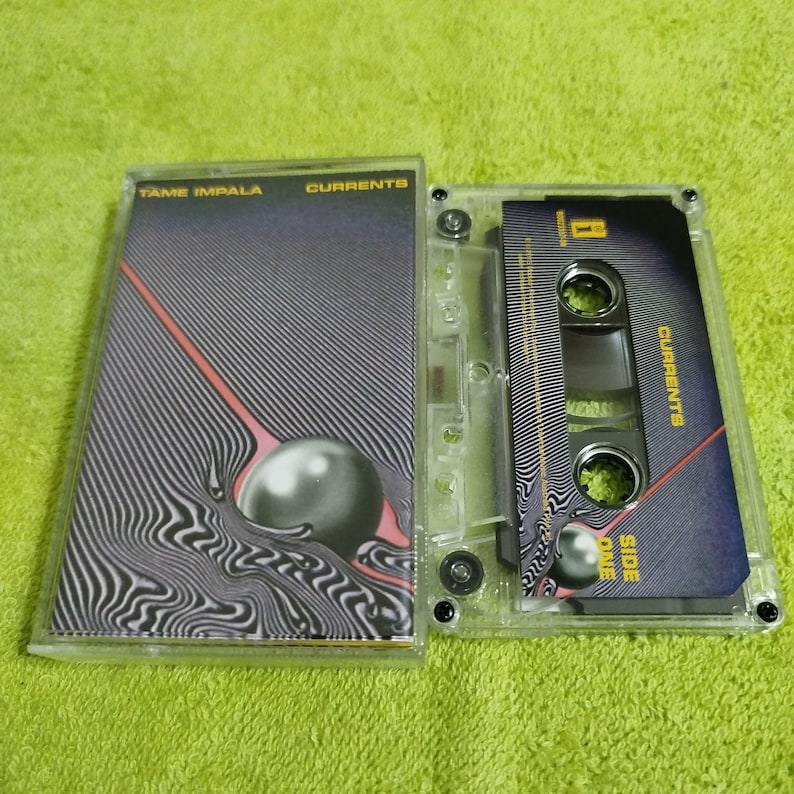 May include: A transparent cassette tape case featuring the album artwork for "Currents" by Tame Impala. The design includes a black and white striped pattern, a pink line, and a grey sphere. The cassette is open.
