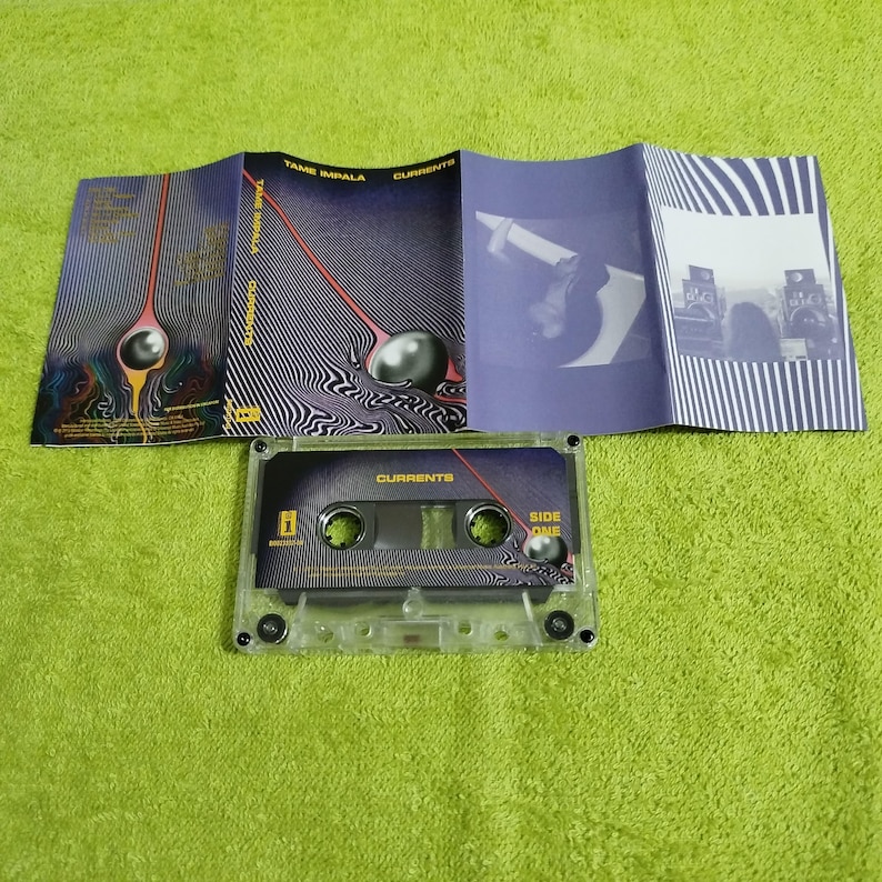 May include: A clear cassette tape labelled "Currents" with a matching album insert. The insert features a black and white striped design with a surreal image of a sphere and a red line. The cassette is on a green surface.