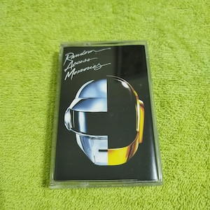 May include: A cassette tape featuring the album art for "Random Access Memories." The cover art is split vertically, with a silver robot helmet on the left and a gold helmet on the right, against a black background. The album title is written in white script.