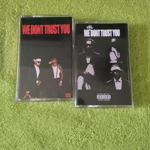 Op de afbeelding: Twee cassettes. Eén heeft de tekst "WE DONT TRUST YOU" in het rood en afbeeldingen van twee mannen. De andere heeft dezelfde tekst en een zwart-wit afbeelding van drie mannen. Beide hebben een waarschuwing voor ouders.