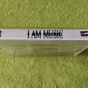 May include: A clear cassette tape with the text "I AM MUSIC" in black. The cassette tape is in a clear plastic case. The background is a green, textured surface.