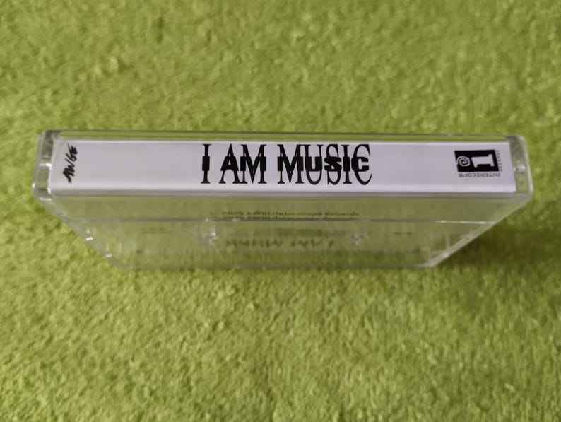May include: A clear cassette tape with the text "I AM MUSIC" in black. The cassette tape is in a clear plastic case. The background is a green, textured surface.
