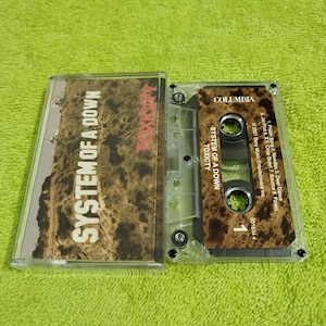 May include: A cassette tape of the album Toxicity by System of a Down. The cassette tape is in a clear plastic case with a black label. The label has the band's name, album title, and track list.