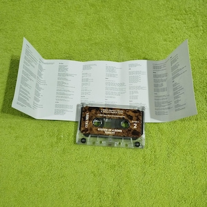 May include: A cassette tape with a brown and black design. The tape is labeled "System of a Down" and "Toxicity". The tape is in a clear plastic case.