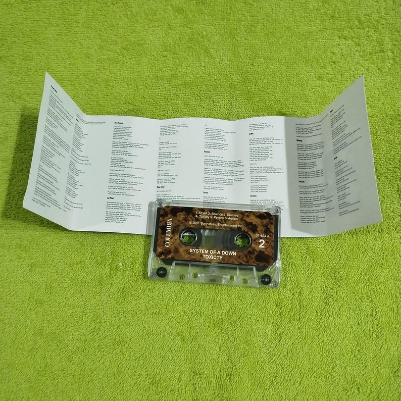 May include: A cassette tape with a brown and black design. The tape is labeled "System of a Down" and "Toxicity". The tape is in a clear plastic case.