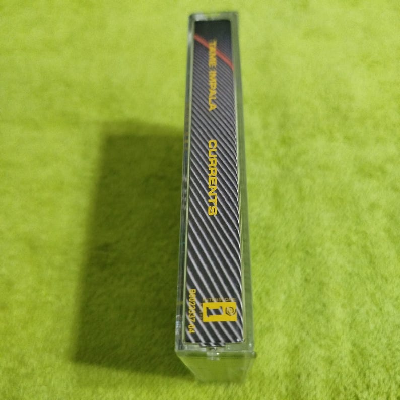 May include: A clear cassette tape case with a black and white striped design. The text "TAME IMPALA CURRENTS" is visible in red and yellow. The case is on a green surface.