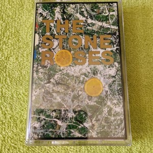 Puede incluir: Una cinta de cassette con la portada del álbum "The Stone Roses". La cubierta presenta un diseño abstracto en blanco, verde y negro, con el nombre de la banda en letras doradas y dos rodajas de limón amarillas.
