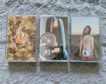 May include: Three cassette tapes with different images. The first tape has a hand giving the middle finger, the second tape has a car, and the third tape has a man with long hair and a bloody bandage on his chest.