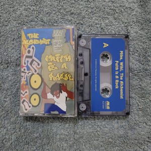May include: A cassette tape with a blue and white label. The label says "Mike, Wiki, The Alchemist Faith Is A Rock" and "ALC Records. All rights reserved. ALC-5051". The cassette tape is in a clear plastic case.