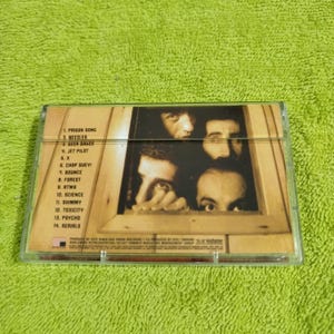 May include: A cassette tape case with a band's photo on the back. The photo shows four men looking out of a window. The band's name is not visible. The track list on the back of the case includes: Prison Song, Needles, Ocean Dance, Jet Pilot, X, Chop Suey!, Bounce, Forest, ATWA, Science, Shimmy, Toxicity, Psycho, and Reruns.
