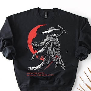 May include: Black sweatshirt featuring a white and red graphic of a witch with a large hat and a red crescent moon in the background. The text "RANNI THE WITCH QUEEN OF THE DARK MOON" is printed below the graphic.