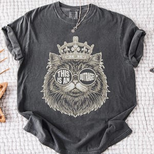 May include: Dark gray t-shirt featuring a detailed illustration of a cat wearing a crown and sunglasses. The sunglasses display the words "THIS IS AN OUTRAGE!". The shirt has rolled-up sleeves and a crew neck.