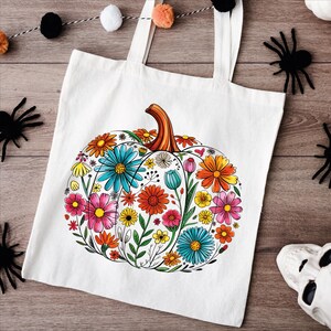 May include: White tote bag featuring a colorful floral pumpkin design. The pumpkin is filled with blue, orange, pink, and yellow flowers, with a brown stem. The bag has two white handles and is made of a sturdy fabric.