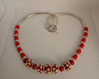 May include: A red and silver beaded necklace with a silver clasp. The necklace has a row of larger red beads with white floral designs.
