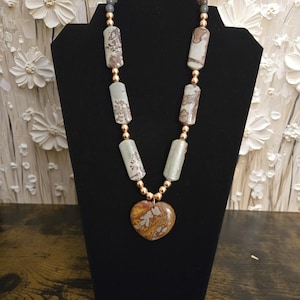 May include: A necklace with a heart-shaped pendant and cylindrical beads. The pendant is a mix of brown, yellow, and white, while the beads are gray and white with brown patterns. Gold-colored beads separate the larger beads.