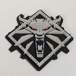 May include: A black and silver emblem featuring a wolf's head with red eyes and an open mouth, set within a diamond-shaped frame. The design includes angular shapes and stylized text around the edges.