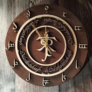 May include: A brown wooden clock with a gold-colored clock face. The clock face features the inscription "One Ring to rule them all, One Ring to find them, One Ring to bring them all and in the darkness bind them" in Elvish script. The clock hands are gold-colored.
