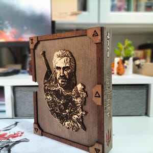 Könnte beinhalten: Holzkiste mit einem detaillierten Relief eines Mannes mit langen Haaren und einem Schwert, wahrscheinlich aus "The Witcher". Die Kiste hat dunkelbraune Kanten und Eckakzente, mit dem Wort "Witcher" an der Seite.