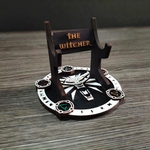Può includere: Un supporto in legno con un cartello che recita "The Witcher" e una base circolare con un'immagine di testa di lupo e rune.