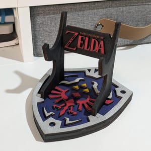 Puede incluir: Un soporte de exhibición de madera oscura con una base en forma de escudo y un diseño de emblema Hyliano en azul, rojo y amarillo. El soporte incluye dos soportes verticales y una placa horizontal con el texto rojo The Legend of Zelda.