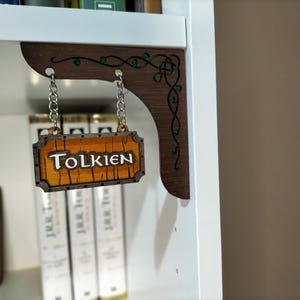 Può includere: Un cartello in legno con la parola "Tolkien" appeso a uno scaffale con un design a vite marrone e verde. Il cartello è appeso a due catene d'argento.