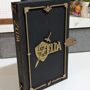 Könnte beinhalten: Eine rechteckige schwarze Holzbox mit goldfarbenen Akzenten. Die Vorderseite zeigt ein erhabenes goldenes Zelda-Logo, einen Schild und ein Schwert-Emblem. Der Buchrücken trägt den Text Zelda Ocarina of Time. Eine kleine beige Lasche mit Niete ist seitlich.