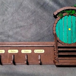 Peut inclure: Un porte-clés en bois en forme de porte de hobbit avec une porte verte et un cadre en bois brun. Le porte-clés a quatre crochets et quatre étiquettes qui indiquent "Frodon", "Bilbon", "Sam" et "Gandalf".