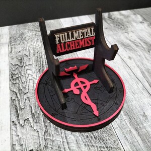 May include: A black wooden stand with a red and black circular base featuring the Fullmetal Alchemist symbol. The stand has a sign that reads "Fullmetal Alchemist".