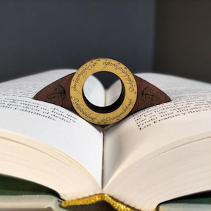 Può includere: Un segnalibro di legno a forma di anello con l'iscrizione "Un anello per domarli tutti, Un anello per trovarli, Un anello per portarli tutti e nell'oscurità legarli" in lingua elfica. Il segnalibro è appoggiato su un libro aperto.