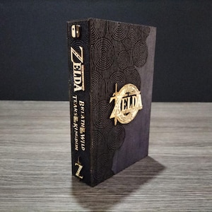 Peut inclure: Boîte décorative sombre avec les mots "ZELDA" et "Breath of the Wild Tears of the Kingdom" en lettres dorées. La boîte présente un emblème doré et un motif stylisé sur sa surface.