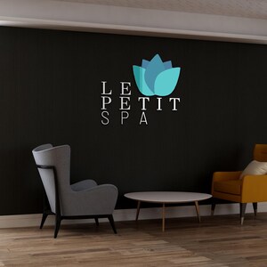 May include: Interior shot of a spa waiting area with a black accent wall. The wall features the text "LE PETIT SPA" and a blue and teal lotus flower logo. Two armchairs, one gray and one yellow, and a round table are in front of the wall.