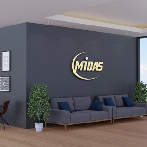 May include: Interior shot of a modern office space with a gray wall featuring a gold "MIDAS" logo. Two gray sofas with blue cushions are in front of the wall. Decorative framed art and plants add to the decor.