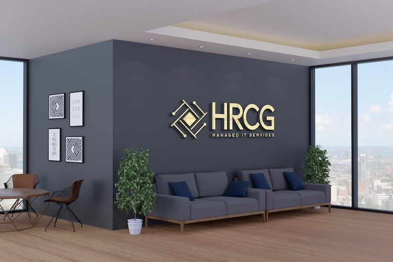 May include: An office interior with a modern design. The room features a dark gray wall with a gold logo that reads "HRCG Managed IT Services." There are two gray sofas with blue cushions, framed artwork, and potted plants. Large windows offer a city view.