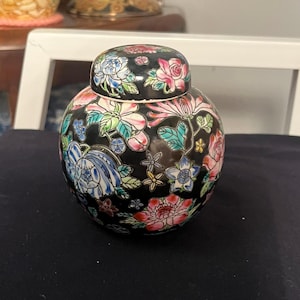 Puede incluir: Un tarro de cerámica negro con tapa, decorado con motivos florales coloridos. El tarro tiene un cuerpo redondo y una parte superior ligeramente acampanada.