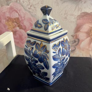 May include: A white ceramic jar with a blue and gold floral design. The jar has a lid with a blue and gold floral design. The jar is a decorative piece.